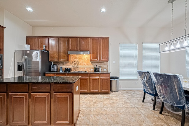 kitchen featuring under cabinet range hood, brown cabinetry, stainless steel fridge, dark stone countertops, and decorative backsplash