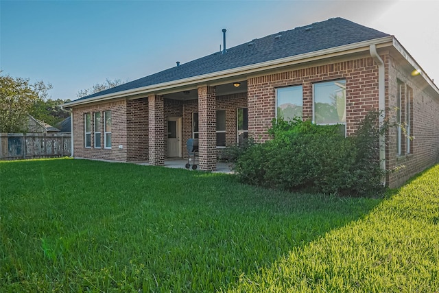 rear view of property with a lawn, brick siding, fence, a patio, and a shingled roof