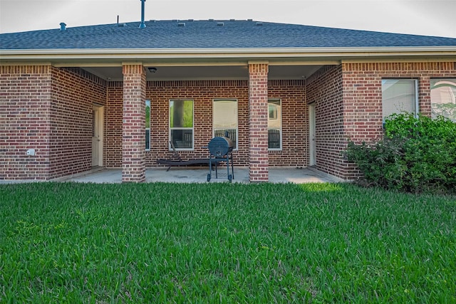 back of house with a lawn, a patio area, roof with shingles, and brick siding