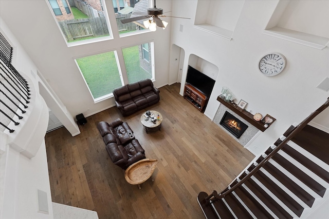 living area featuring a towering ceiling, a warm lit fireplace, a ceiling fan, wood finished floors, and stairs