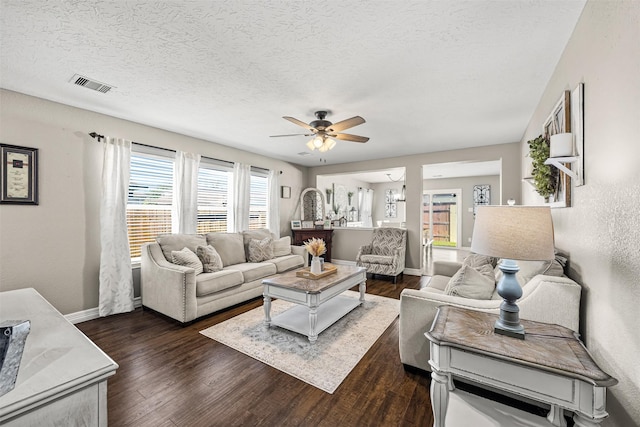 living room with a textured ceiling, visible vents, baseboards, dark wood-type flooring, and ceiling fan