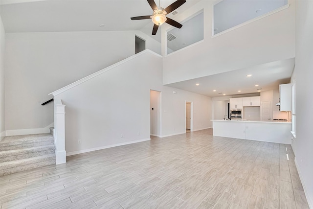 unfurnished living room featuring ceiling fan, stairway, light wood-type flooring, a towering ceiling, and baseboards