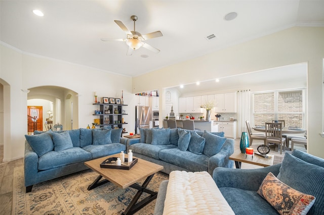 living area with arched walkways, ornamental molding, ceiling fan, vaulted ceiling, and light wood-style floors