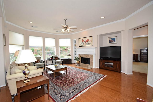 living area with wood finished floors, baseboards, ceiling fan, ornamental molding, and a high end fireplace