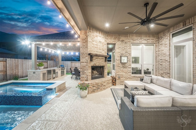 patio terrace at dusk featuring a fenced backyard, an outdoor kitchen, an outdoor living space with a fireplace, and ceiling fan