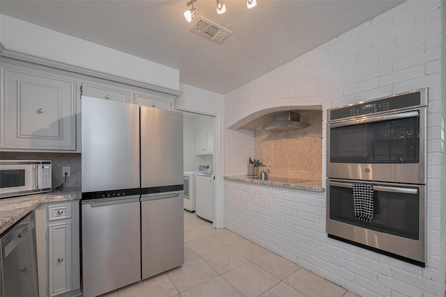 kitchen featuring stainless steel appliances, independent washer and dryer, wall chimney exhaust hood, backsplash, and light tile patterned floors