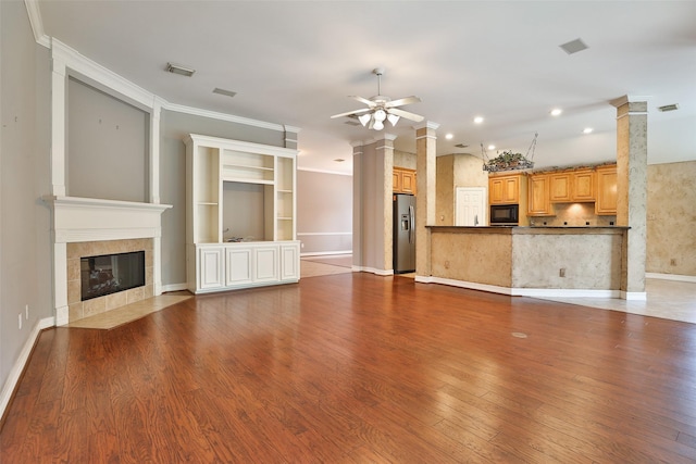 unfurnished living room with a tiled fireplace, ceiling fan, visible vents, decorative columns, and wood finished floors