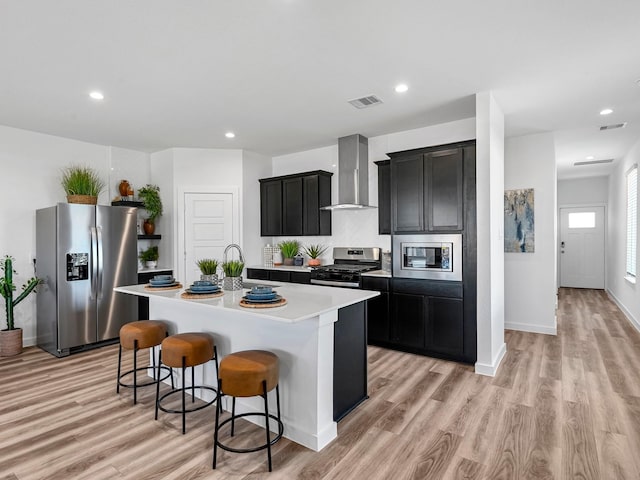 kitchen with dark cabinets, a kitchen breakfast bar, wall chimney exhaust hood, stainless steel appliances, and an island with sink