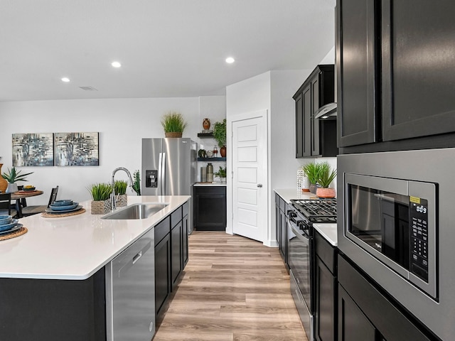 kitchen with light wood finished floors, a sink, stainless steel appliances, a kitchen island with sink, and light countertops