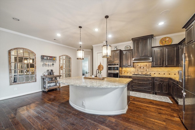 kitchen with appliances with stainless steel finishes, under cabinet range hood, light stone countertops, dark brown cabinets, and an island with sink