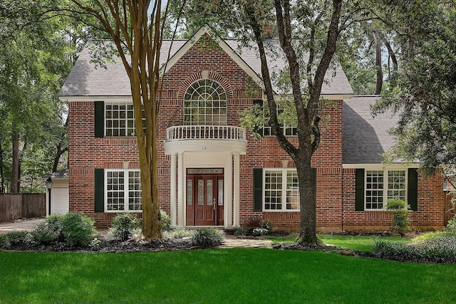 colonial home with a balcony, brick siding, fence, a front yard, and roof with shingles