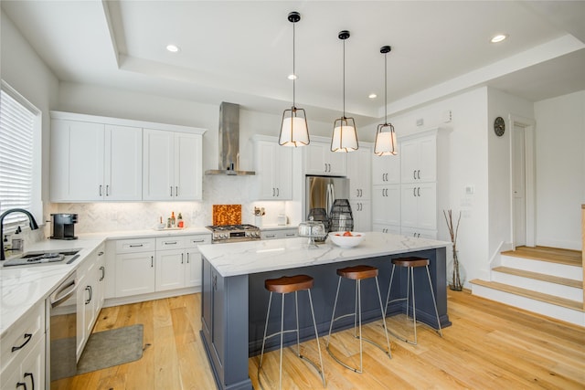 kitchen with wall chimney range hood, a sink, stainless steel appliances, a kitchen island, and light wood finished floors