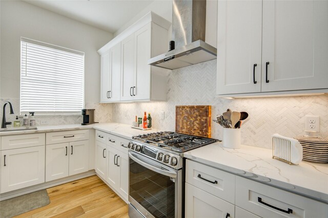 kitchen featuring wall chimney exhaust hood, stainless steel gas range, a sink, light stone countertops, and white cabinets