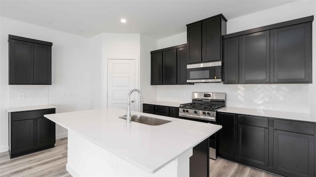 kitchen featuring a sink, light wood-type flooring, a center island with sink, appliances with stainless steel finishes, and decorative backsplash