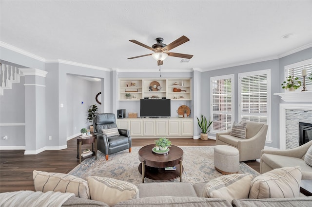 living area with ceiling fan, dark wood finished floors, plenty of natural light, a stone fireplace, and ornamental molding