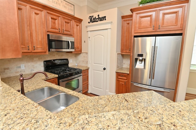 kitchen with a sink, brown cabinetry, crown molding, light stone countertops, and appliances with stainless steel finishes