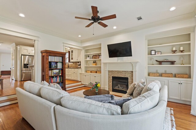 living room featuring ornamental molding, ceiling fan, light wood-style flooring, a stone fireplace, and built in features