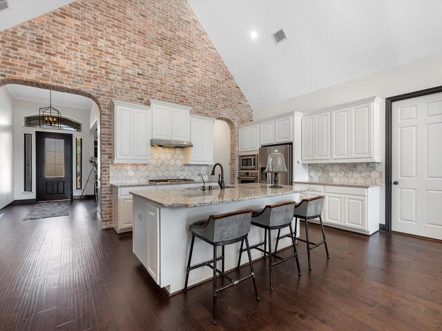 kitchen featuring arched walkways, appliances with stainless steel finishes, high vaulted ceiling, white cabinetry, and a center island with sink