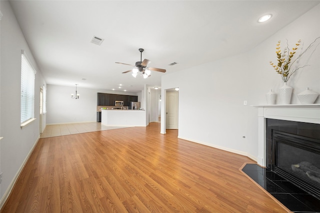unfurnished living room with a tile fireplace, ceiling fan, light wood-style flooring, a chandelier, and baseboards