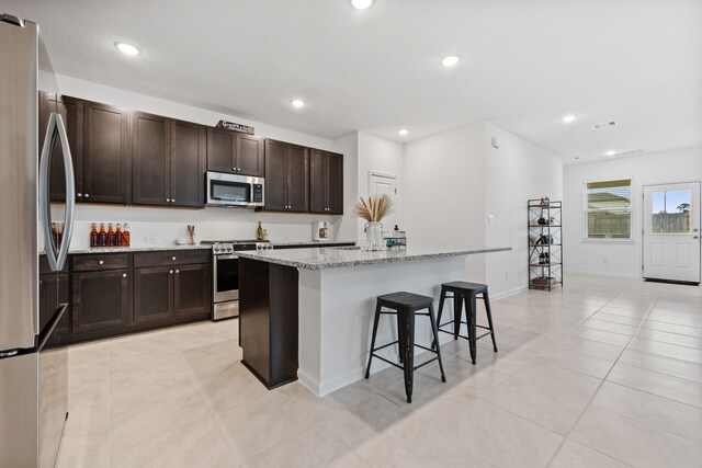 kitchen featuring appliances with stainless steel finishes, a kitchen bar, a kitchen island with sink, light stone counters, and light tile patterned floors