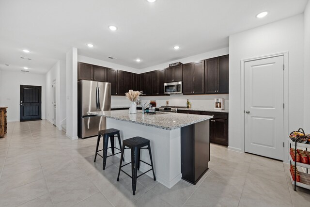 kitchen with stainless steel appliances, a kitchen bar, dark brown cabinets, a kitchen island, and light stone counters