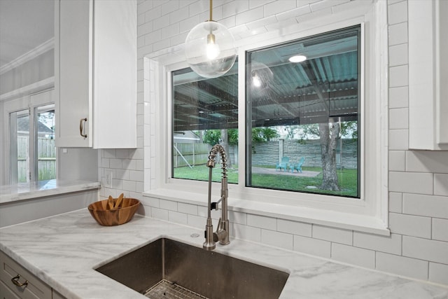 kitchen view of a sink, light stone countertops, ornamental molding, white cabinets, and pendant lighting