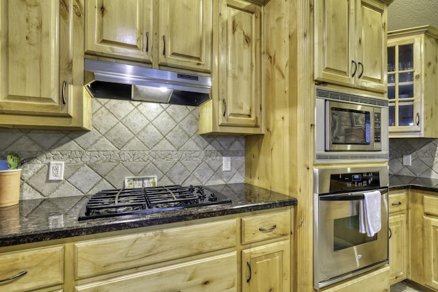 kitchen featuring stainless steel appliances, under cabinet range hood, backsplash, glass insert cabinets, and dark stone countertops