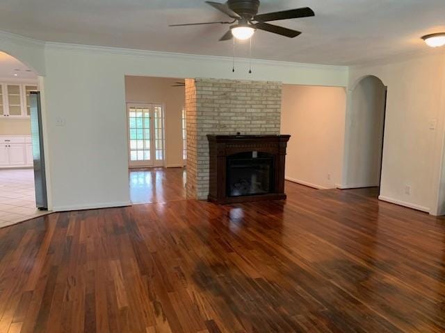 unfurnished living room featuring arched walkways, ceiling fan, wood-type flooring, a brick fireplace, and crown molding