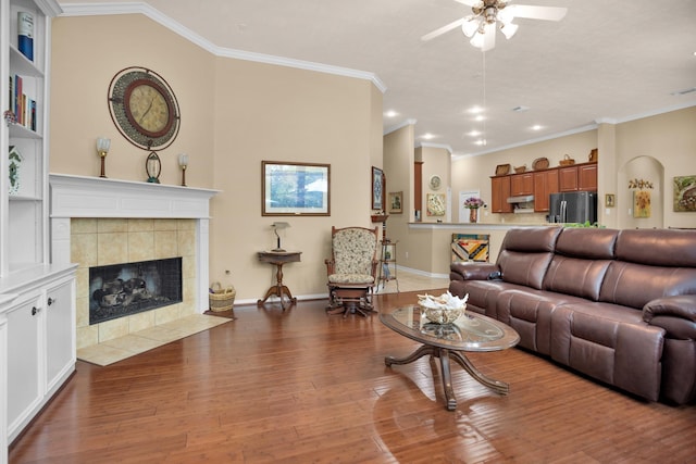 living area featuring ceiling fan, ornamental molding, wood finished floors, a tiled fireplace, and baseboards
