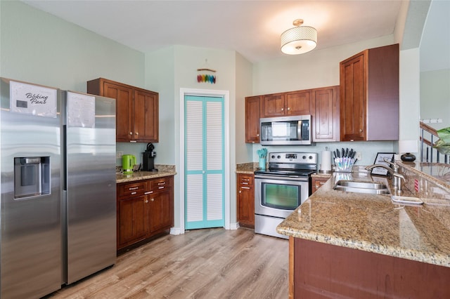 kitchen featuring stainless steel appliances, a sink, brown cabinetry, light stone countertops, and light wood finished floors