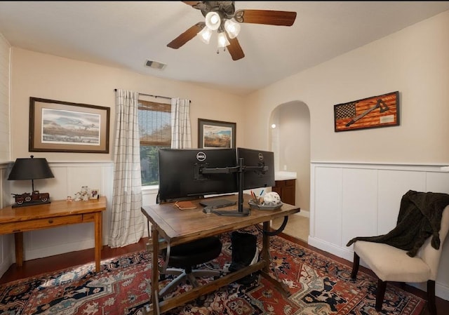 office area featuring arched walkways, a decorative wall, ceiling fan, wood finished floors, and a wainscoted wall