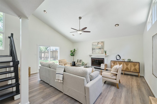 living area with a ceiling fan, stairway, light wood-style flooring, a glass covered fireplace, and high vaulted ceiling