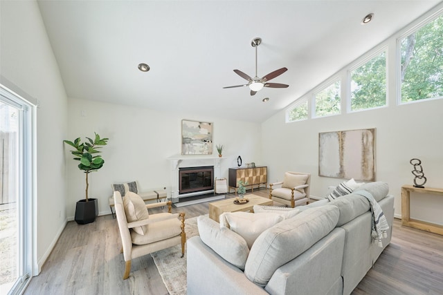 living room featuring a ceiling fan, high vaulted ceiling, light wood-style flooring, a glass covered fireplace, and baseboards