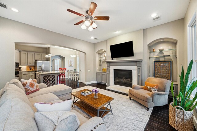 living area featuring arched walkways, ceiling fan, dark wood-type flooring, a glass covered fireplace, and built in features