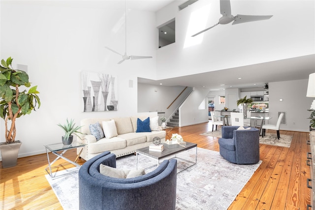 living area featuring a ceiling fan, stairs, light wood-type flooring, baseboards, and a towering ceiling