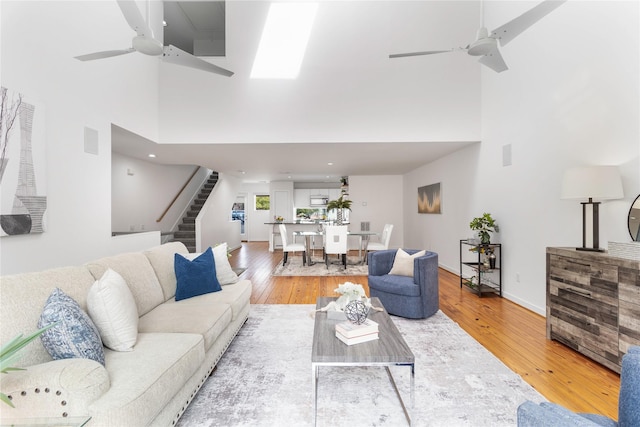 living room featuring ceiling fan, stairs, a high ceiling, light wood-style flooring, and baseboards