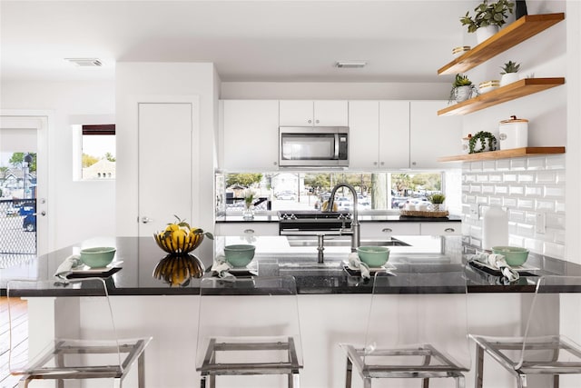 kitchen with stainless steel microwave, open shelves, dark countertops, white cabinets, and healthy amount of natural light