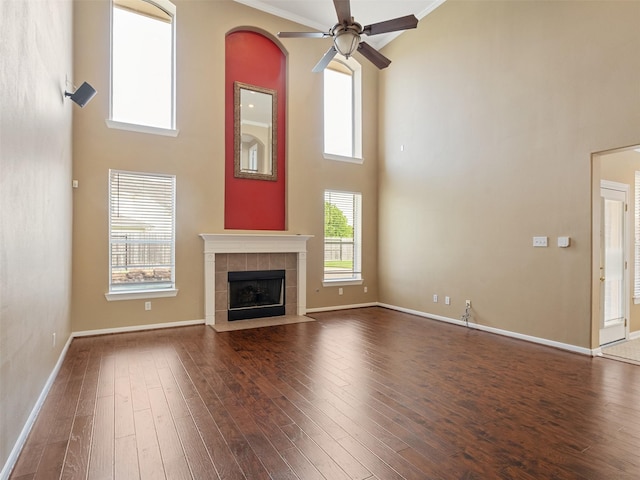 unfurnished living room with a high ceiling, a ceiling fan, a tile fireplace, dark wood-type flooring, and baseboards