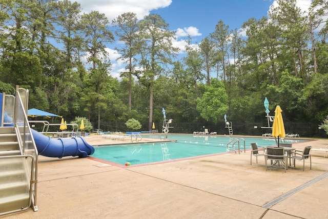 community pool featuring a water slide, a patio area, fence, and view of scattered trees
