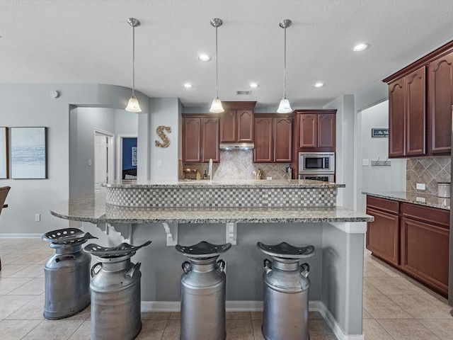 kitchen featuring stainless steel microwave, backsplash, a kitchen breakfast bar, light stone countertops, and hanging light fixtures