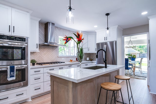 kitchen featuring wall chimney range hood, appliances with stainless steel finishes, a sink, backsplash, and light wood finished floors