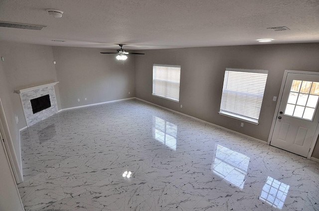 unfurnished living room featuring plenty of natural light, ceiling fan, a textured ceiling, a fireplace, and light marble finish floors