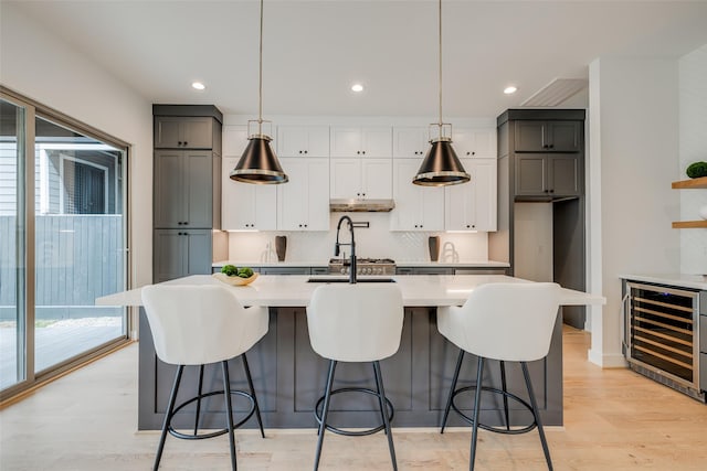 kitchen with wine cooler, light countertops, under cabinet range hood, light wood-type flooring, and tasteful backsplash