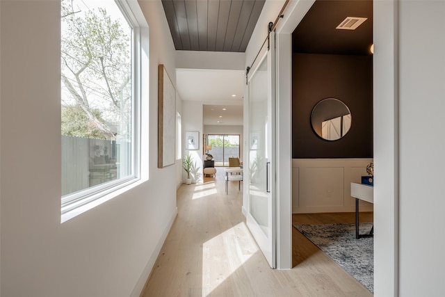 hall with a barn door, light wood finished floors, a wainscoted wall, and a decorative wall