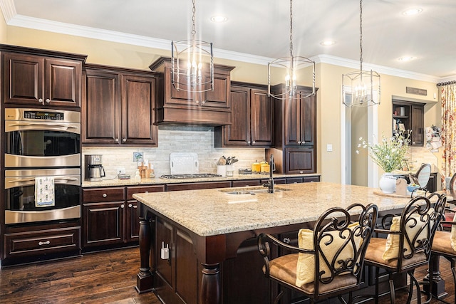 kitchen with appliances with stainless steel finishes, a chandelier, a sink, a center island with sink, and light stone countertops