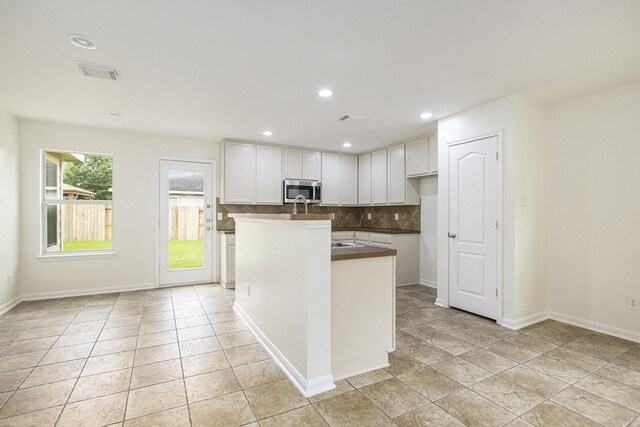kitchen with stainless steel microwave, white cabinetry, decorative backsplash, a center island with sink, and baseboards