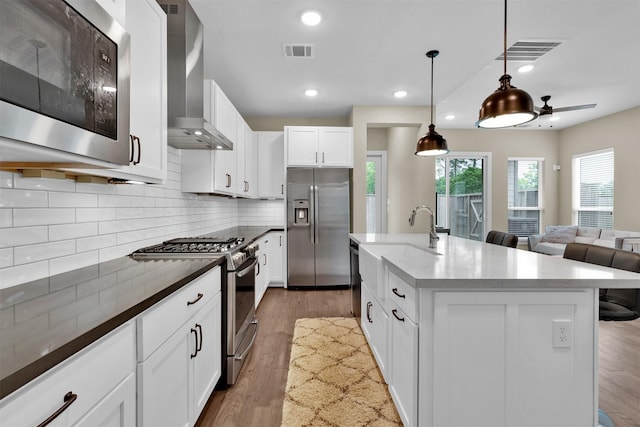 kitchen featuring stainless steel appliances, wall chimney range hood, decorative backsplash, light wood-style flooring, and decorative light fixtures
