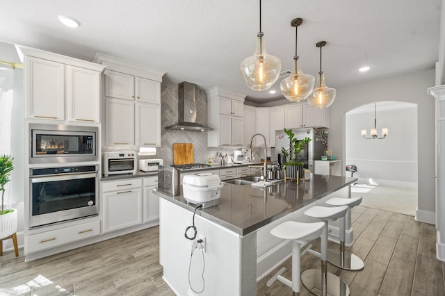 2723 Camellia Ave, Brookshire, TX 77423 - kitchen featuring stainless steel appliances, wall chimney exhaust hood, dark countertops, arched walkways, and light wood-style flooring