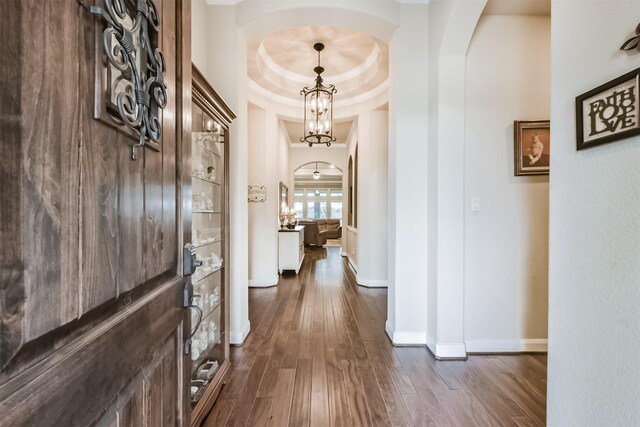 hall with arched walkways, dark wood finished floors, baseboards, and a raised ceiling