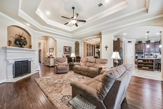 living area featuring a ceiling fan, arched walkways, a tray ceiling, a fireplace with raised hearth, and light wood finished floors